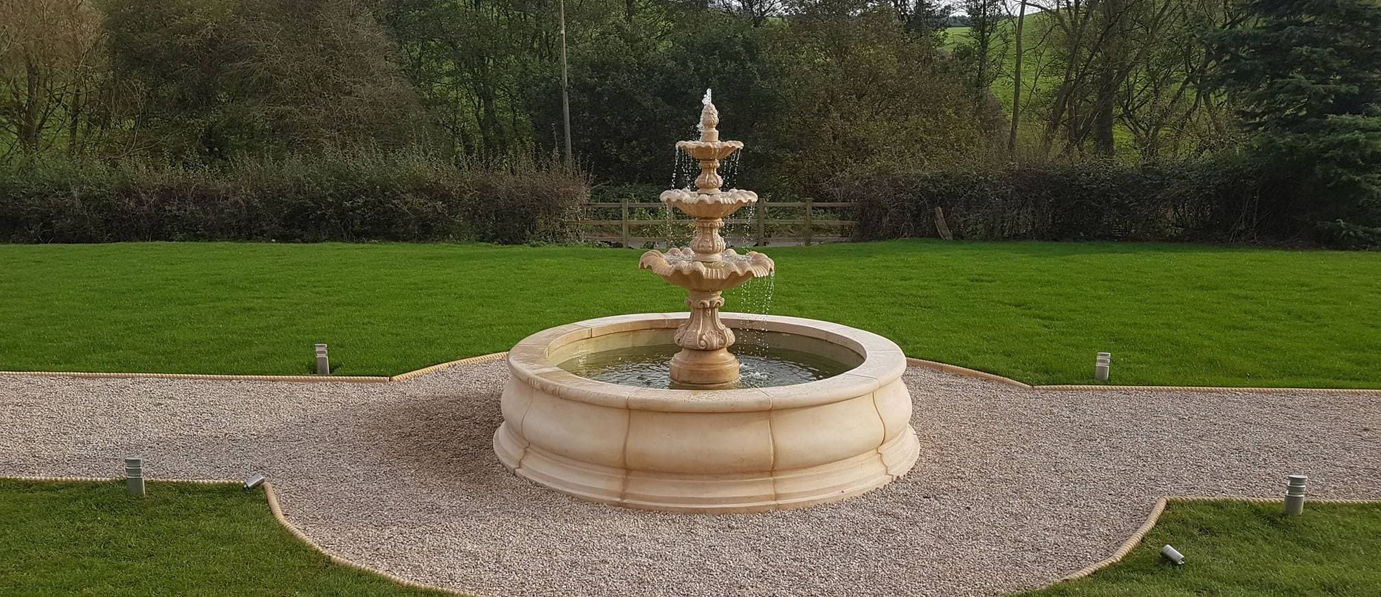 A tiered stone fountain surrounded by trimmed hedges in a landscaped garden